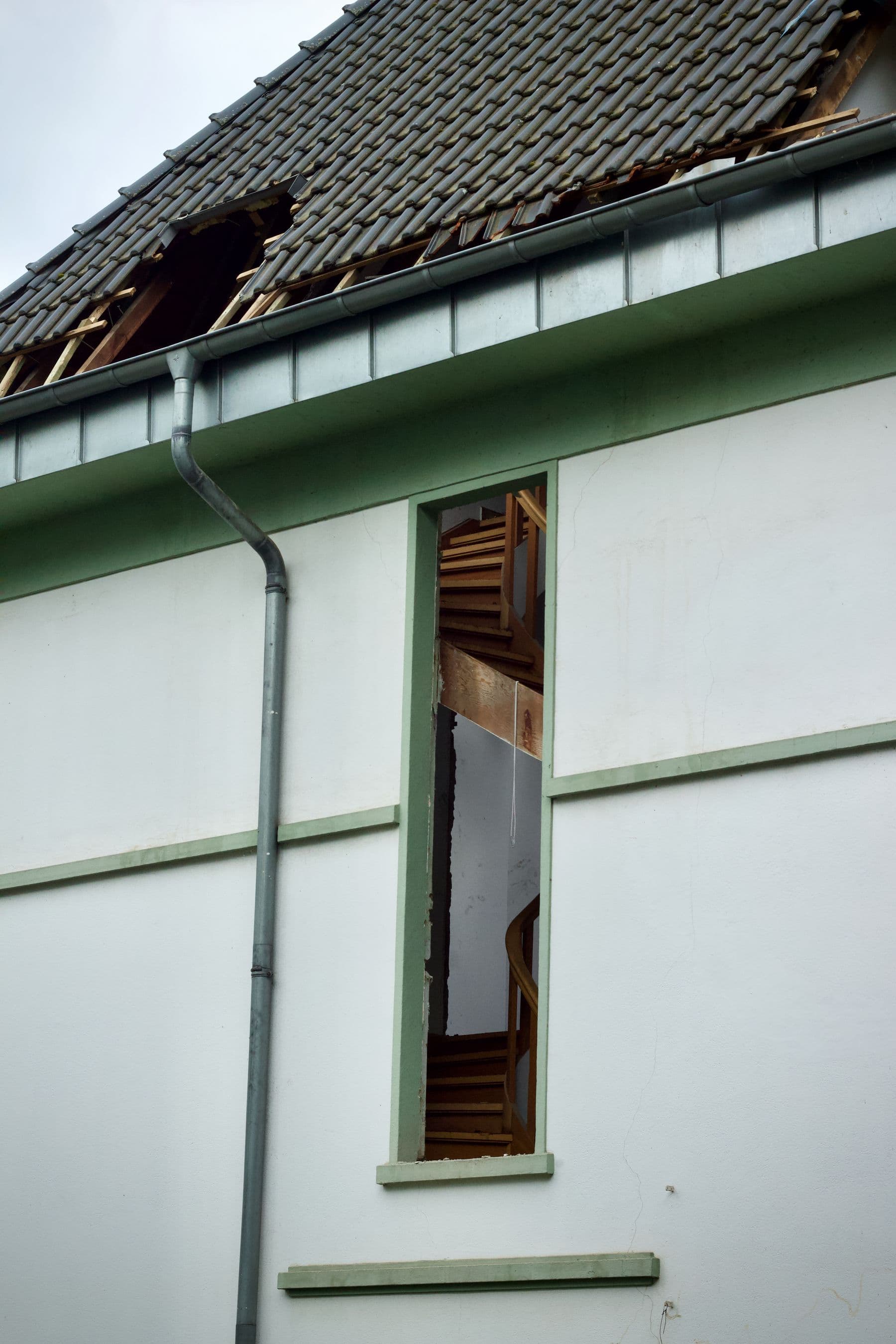 A white house wall with a broken tile roof above it and an empty window opening that shows a wooden staircase inside.