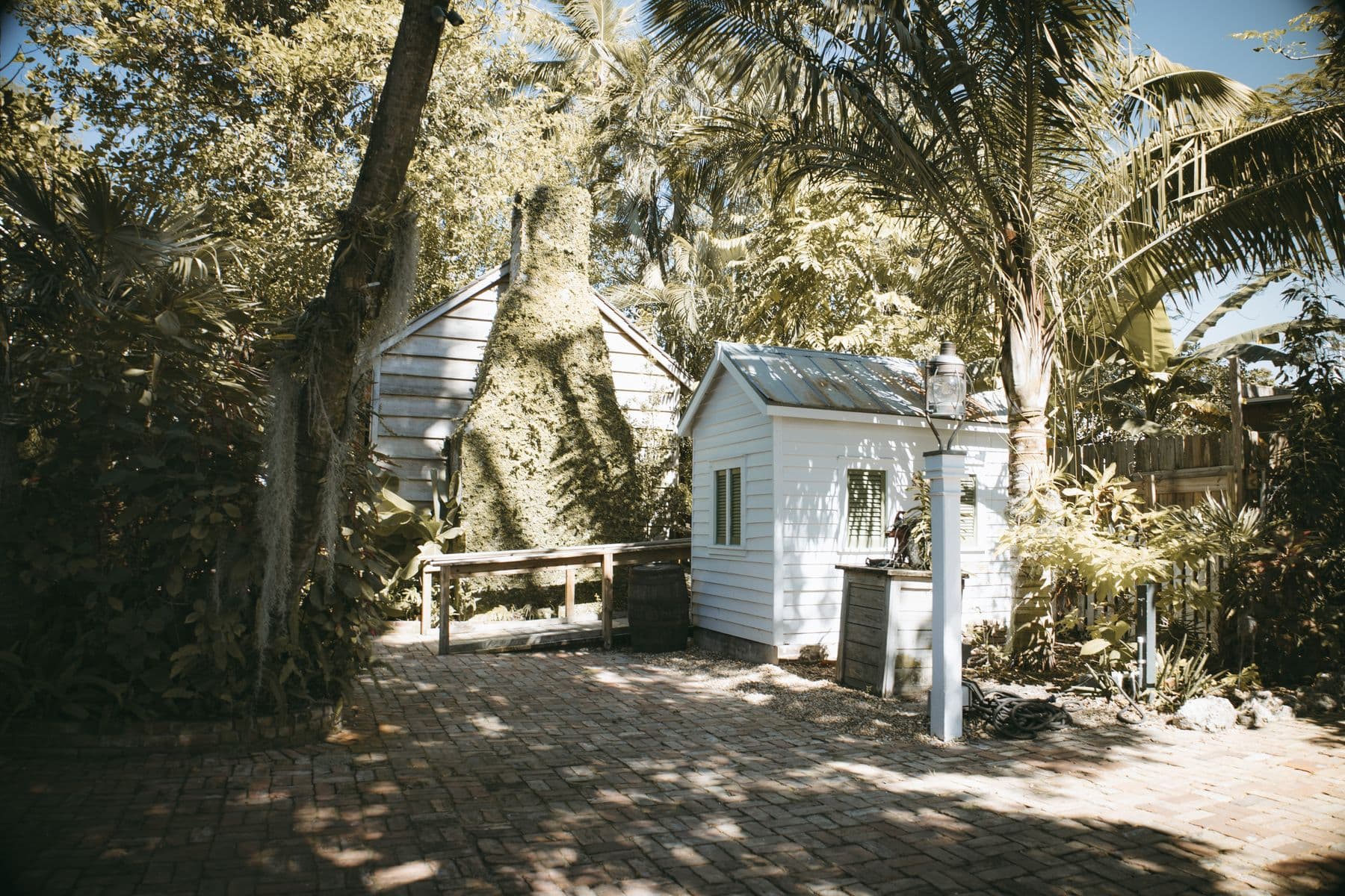 A small white shed and an old wooden building among tropical trees on a brick-paved yard in Key West.