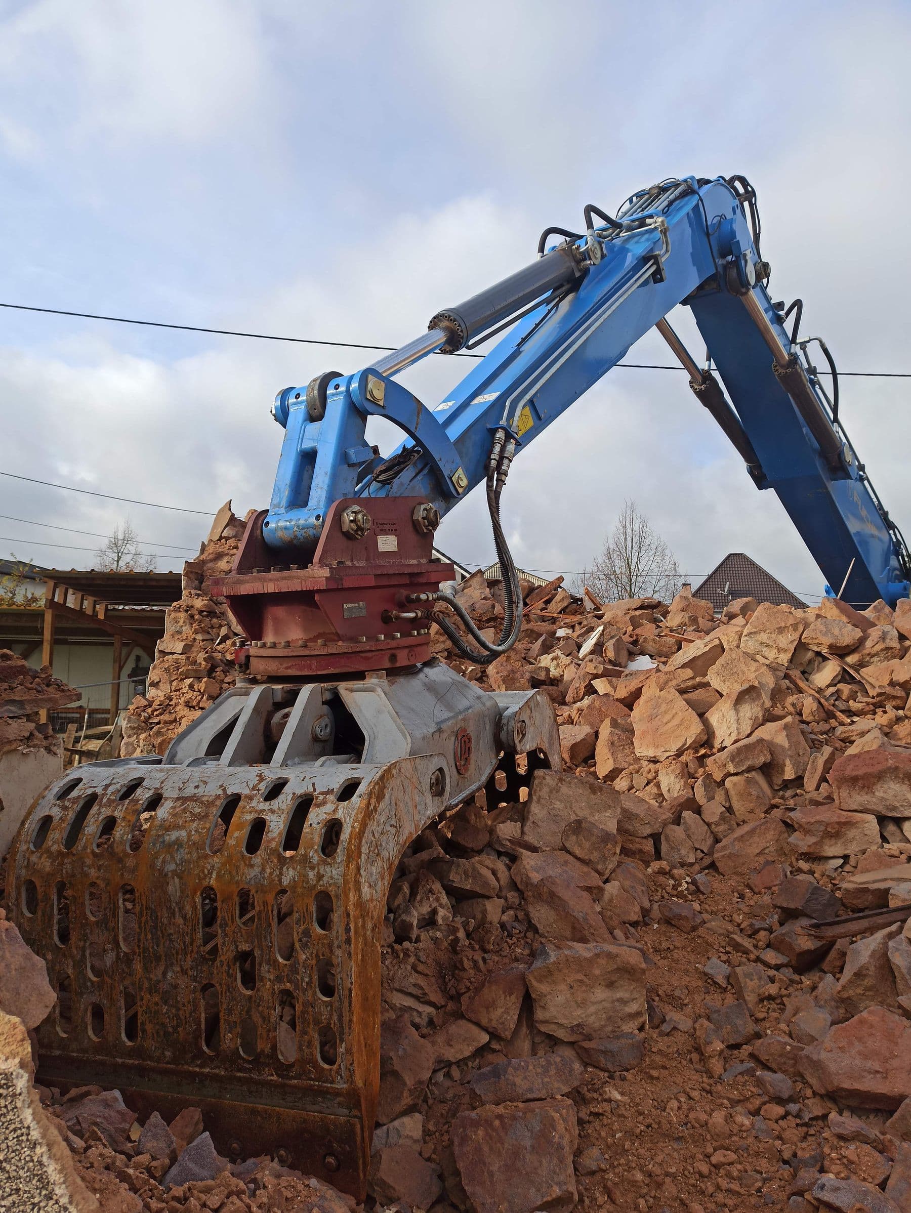 A blue excavator with a rusty grapple bucket resting on a pile of broken stone and rubble.
