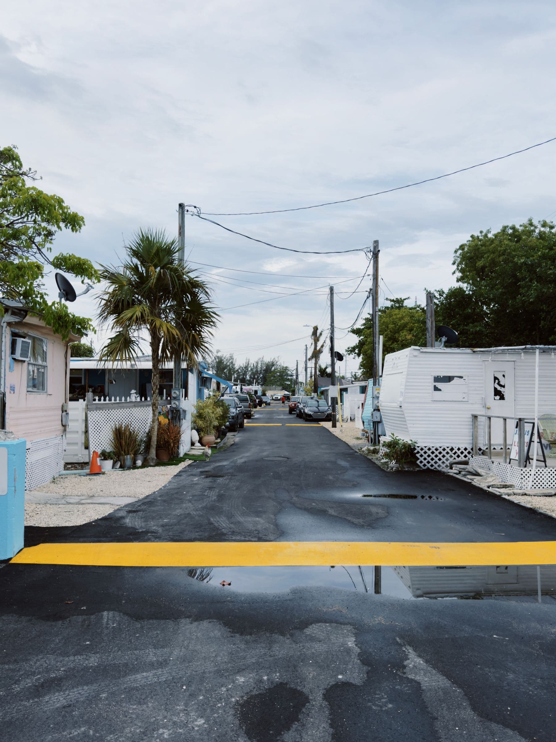 A narrow paved street through a mobile home park lined with trailers and palm trees under a cloudy sky.